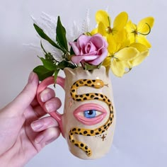 a hand holding a vase with flowers in it and a face painted on the side