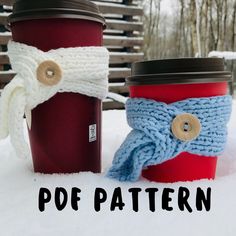 two knitted coffee cups sitting in the snow, one with a scarf on it