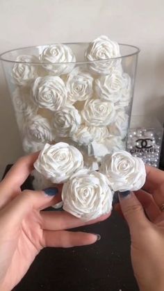 two hands are holding small white roses in a glass vase on a table next to other items