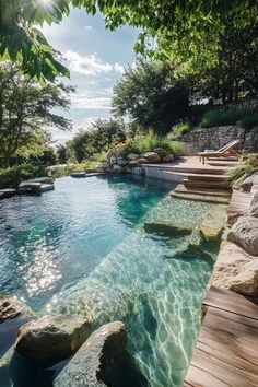 an outdoor swimming pool surrounded by rocks and water features clear blue water, green trees, and sun shining through the clouds