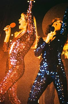 two women in sequin outfits singing on stage with microphones up to their ears