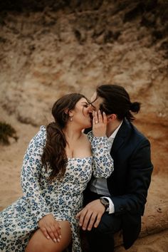 a man and woman sitting on the ground kissing in front of a rocky mountain side