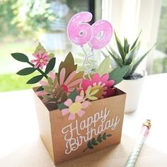 a birthday card in a box on a table with a pen and flower pot next to it