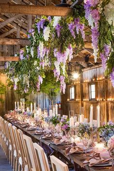 a long table with purple flowers hanging from it's ceiling and candles on the tables