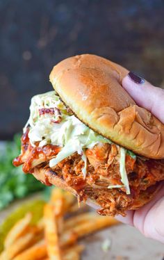 a hand holding up a pulled pork sandwich with coleslaw on it and french fries in the background