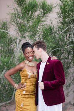 a man and woman standing next to each other in front of some bushes with their arms around each other