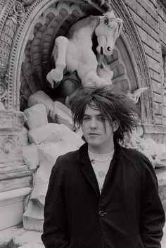 black and white photograph of a man with long hair standing in front of a horse statue