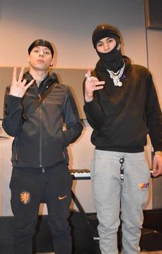 two young men standing next to each other in front of a white wall wearing black clothing