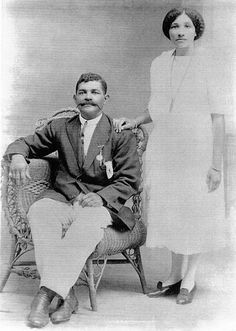 an old black and white photo of two people posing for the camera, one is sitting on a chair