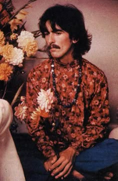 a man with long hair sitting next to a vase filled with flowers and wearing a beaded necklace