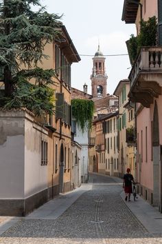 an old cobblestone street with two people walking down one side and a clock tower in the background