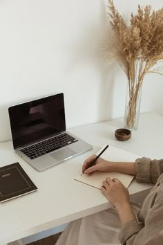 a person sitting at a desk with a laptop and notebook in front of them, writing on a notepad