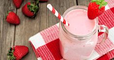 a strawberry smoothie in a mason jar with strawberries scattered around it on a red and white checkered napkin