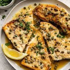 fish with capers and lemon on a white plate next to a bowl of sauce