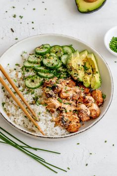 a white bowl filled with rice, cucumber and chicken next to chopsticks