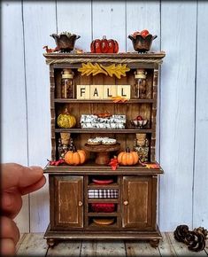 a hand holding a small doll house with fall decorations on the top and bottom shelf