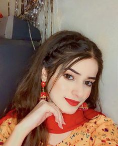 a woman with long brown hair wearing red and gold earrings, sitting on a couch