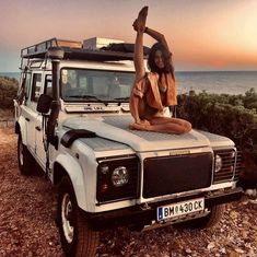 a woman is sitting on top of a vehicle by the ocean with her arms in the air