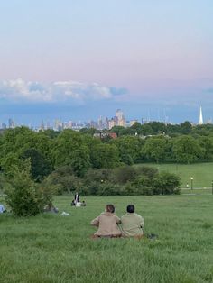 two people sitting on the grass flying a kite in a park with trees and buildings in the background