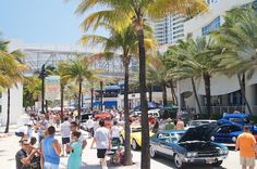 a crowd of people walking down a street next to tall palm trees and parked cars