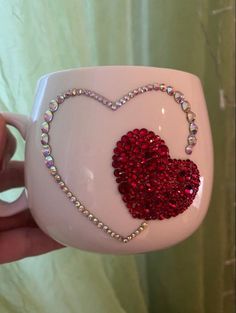 a hand holding a coffee mug with a red heart on the front and beaded chain attached to it