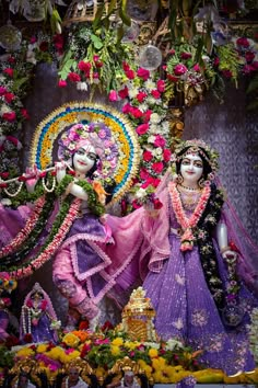 two people dressed in purple and pink are standing next to each other with flowers on the wall behind them