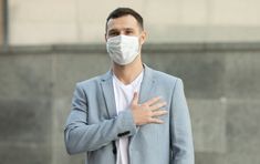 a man wearing a face mask standing in front of a building with his hands on his chest