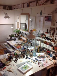 a cluttered desk with lots of art supplies on it and a lamp hanging from the ceiling