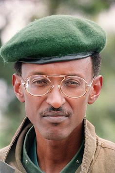 a man with glasses and a green hat is looking at the camera while standing in front of trees