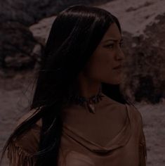 a woman with long black hair wearing a brown shirt and fringed necklace standing in front of some rocks