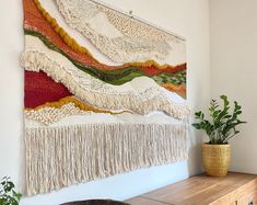 a wall hanging with fringes and plants on top of a wooden table next to a potted plant