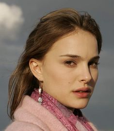 a close up of a person wearing a pink sweater and earrings with clouds in the background