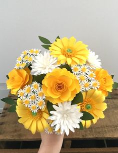 a hand holding a bouquet of yellow and white flowers on top of a wooden table