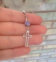 a person is holding a small cross belly ring with a purple crystal bead on it