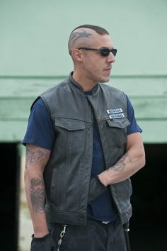 a man with tattoos on his arm wearing a leather vest and black sunglasses standing in front of a green building