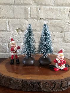 three small figurines sitting on top of a piece of wood next to trees