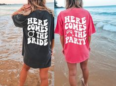 two women standing on the beach wearing matching shirts that say here comes the bride and here comes the party