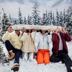 three people sitting in the snow holding up a surfboard above their heads with trees behind them