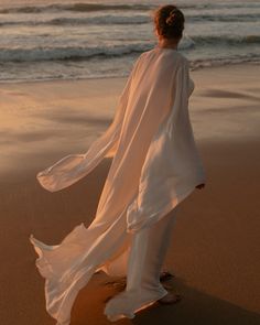 a woman standing on top of a sandy beach next to the ocean with her dress blowing in the wind