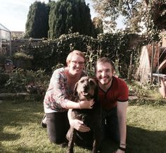 two people kneeling down with a dog in their lap on the grass near some bushes