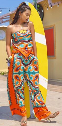 a woman standing next to a surfboard wearing an orange and blue patterned jumpsuit