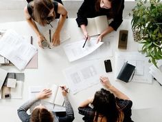 three women are sitting at a table with books and notebooks on it, surrounded by papers