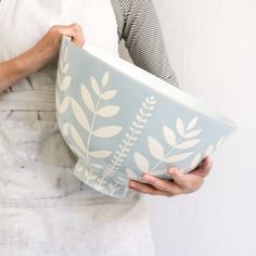 a woman is holding a blue bowl with white leaves on it and she has an apron over her shoulder