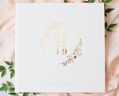 a wedding guest book with gold foil on it and greenery around the edges, surrounded by pink flowers