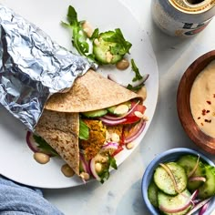 a white plate topped with a wrap and salad next to a bowl of soup on top of a table