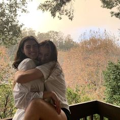 two young women hugging each other on a wooden deck in front of trees and hills