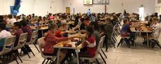 a large group of people sitting at tables eating food in a room with white walls