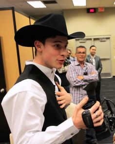 a young man wearing a black cowboy hat and holding a camera in front of other men