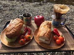 two plates with sandwiches, tomatoes and an apple on a picnic table next to water