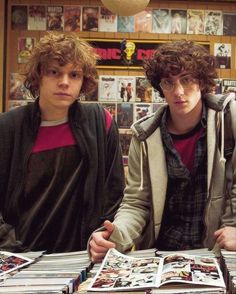 two young men sitting at a table covered in comic books, one with curly hair and the other without